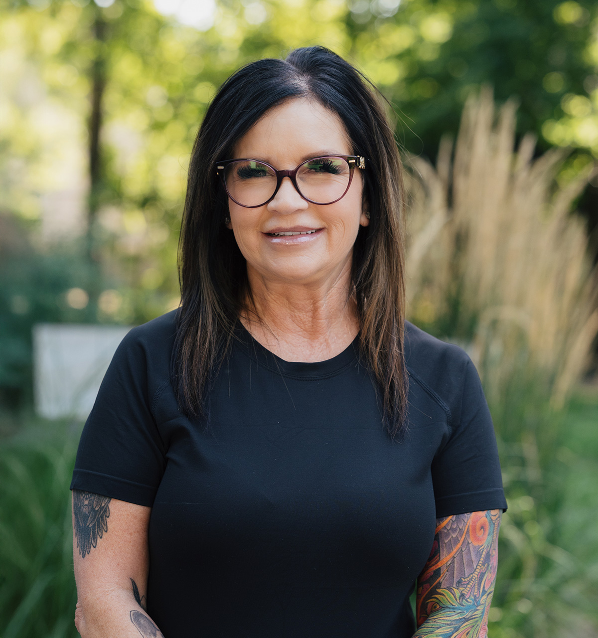 Smiling woman with dark hair, glasses, and colorful tattoos on her arms wearing a black shirt outdoors.