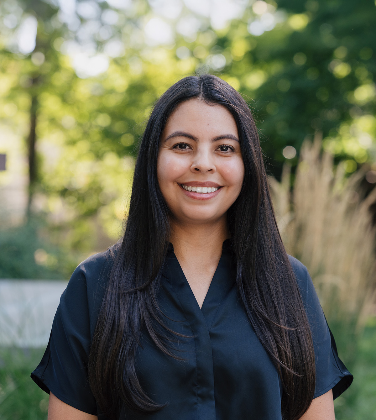 Smiling woman with long dark hair wearing a black shirt outdoors with blurred greenery in the background.
