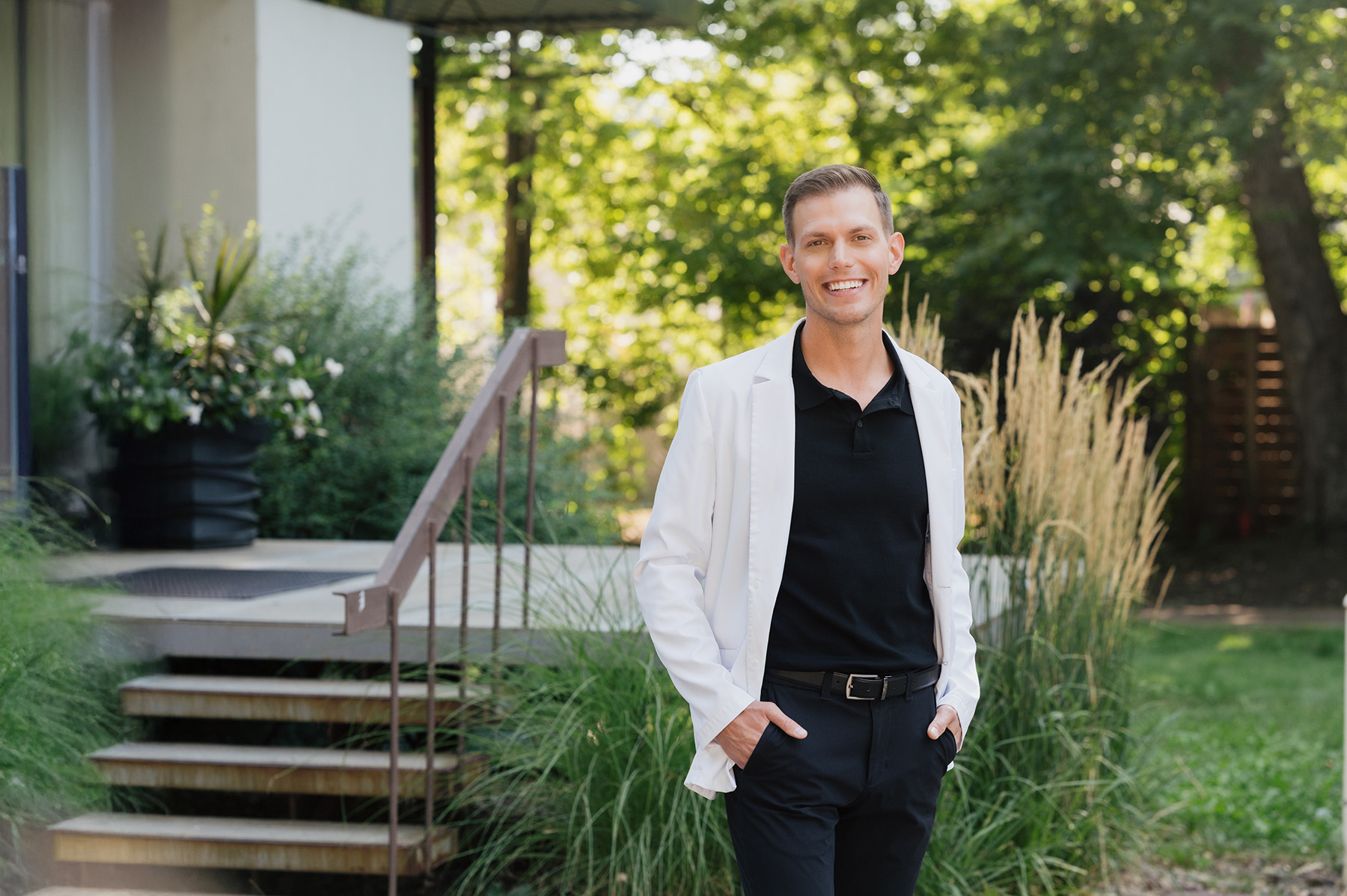 Smiling man wearing a white lab coat and black polo shirt standing outdoors with hands in pockets.
