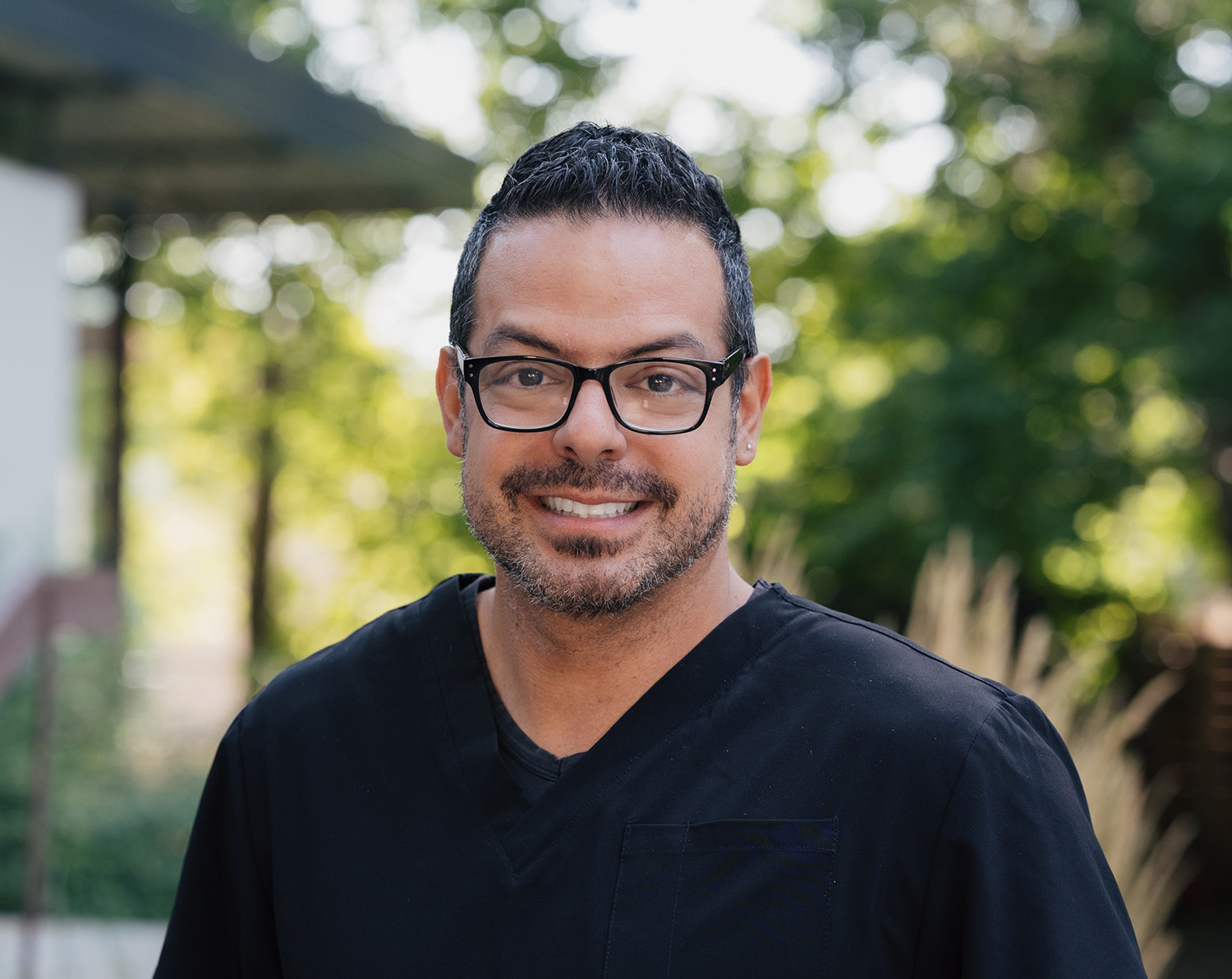 Smiling man with glasses and short curly hair wearing a black scrub top outdoors.