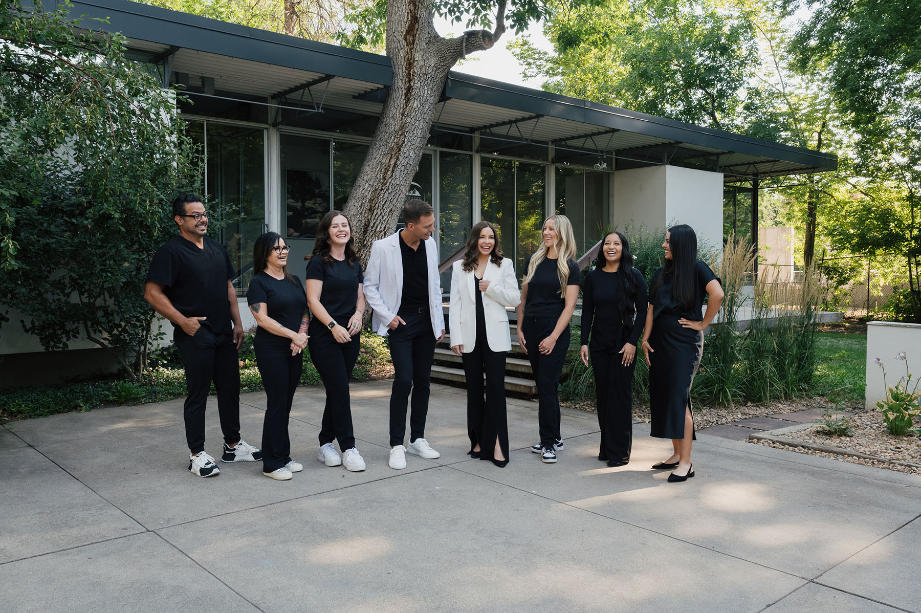 Group of seven men and women standing outdoors on concrete, smiling and chatting in front of a modern building and trees.
