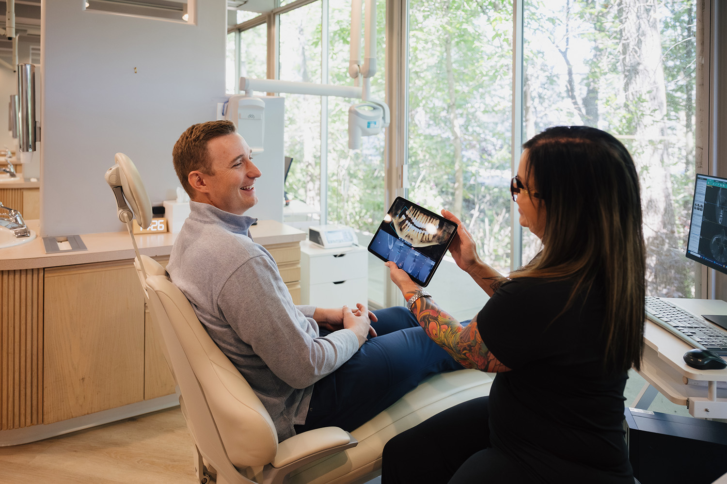 Dentist with tattoos showing a dental X-ray on a tablet to a male patient seated in a dental chair.