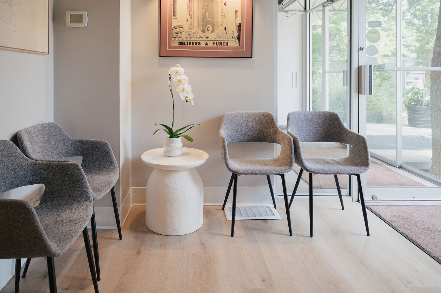 Minimalist waiting area with four gray upholstered chairs around a white round table holding a white orchid, near a glass door entrance.