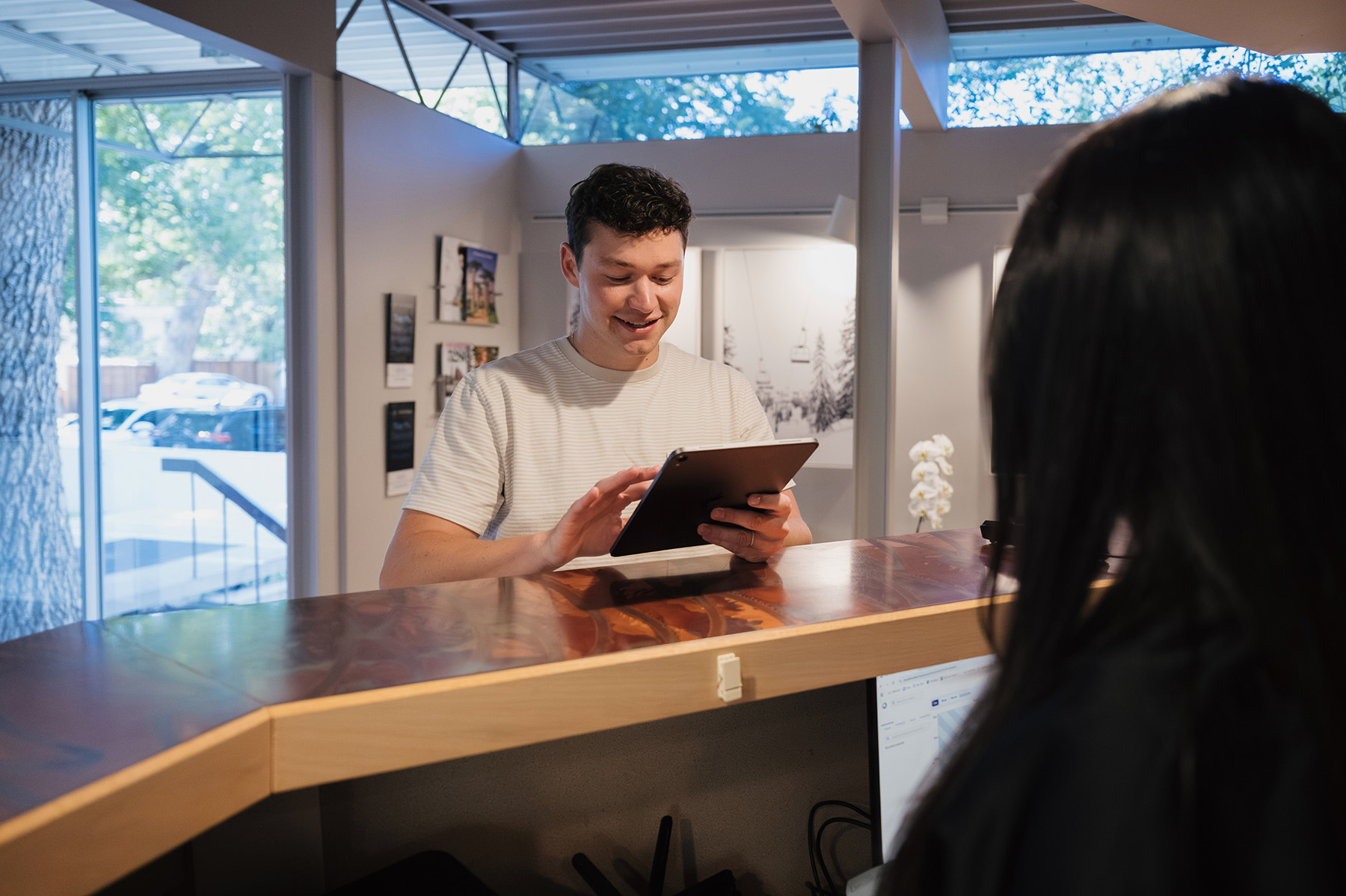 Smiling man in a striped t-shirt using a tablet at a reception desk with a woman sitting nearby.