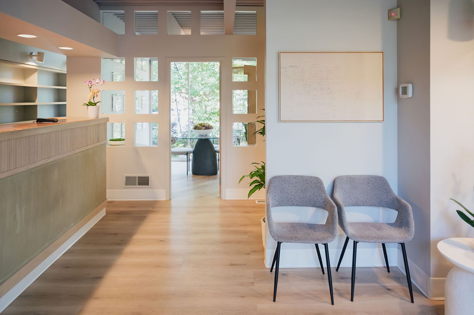 Modern waiting area with two grey chairs, a reception desk, and a doorway leading to a room with a glass table and chairs.