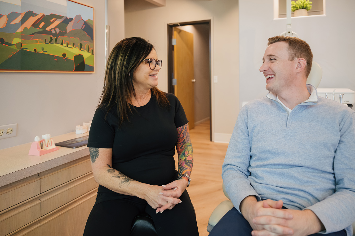 Woman with tattoos and glasses talking and smiling with a man in a dental office.