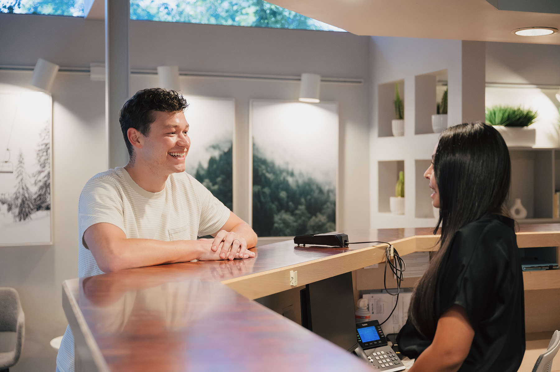 Smiling man leaning on a wooden reception desk talking to a seated woman with long black hair.