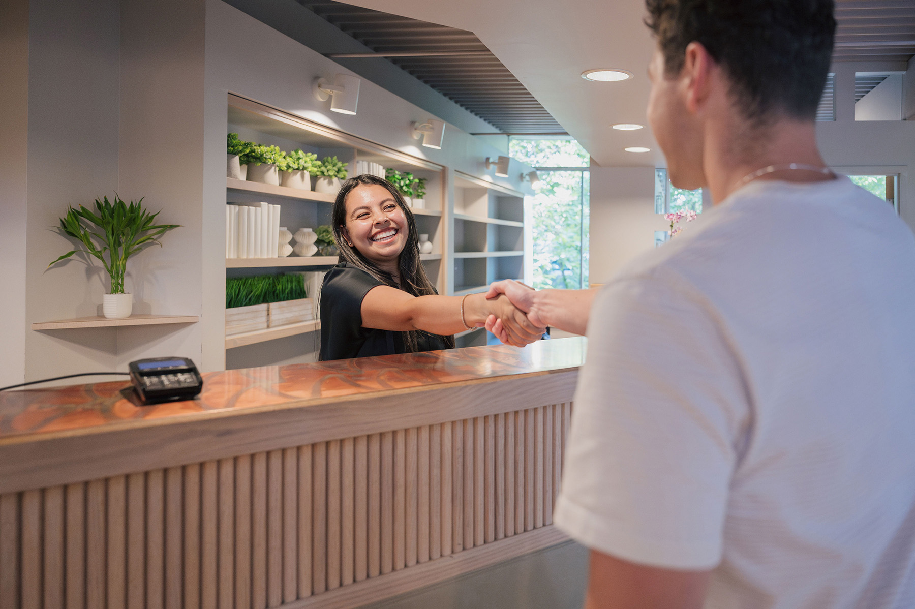 Smiling receptionist shaking hands with a man across a wooden front desk in a modern office.