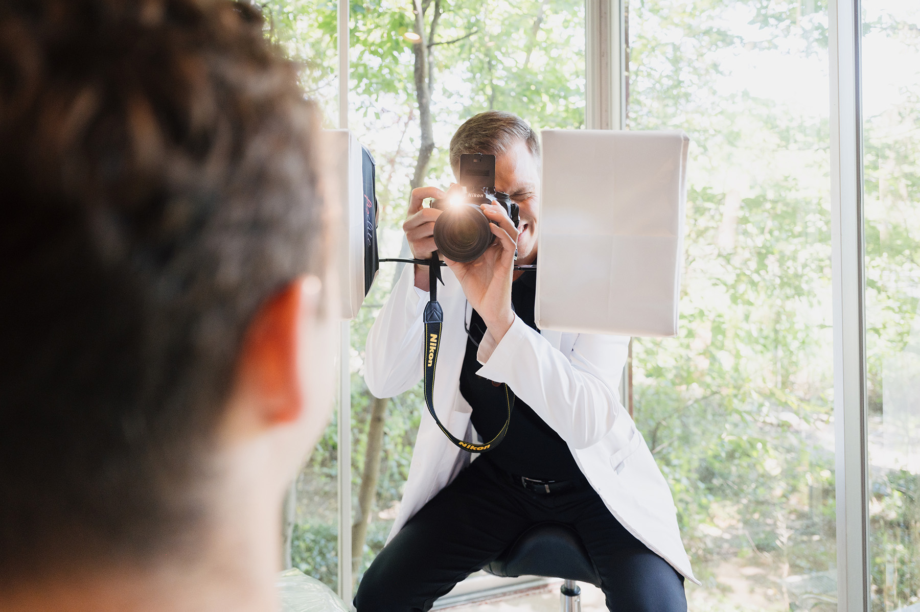 Photographer in a white coat taking a photo with a Nikon camera in a bright room with large windows.