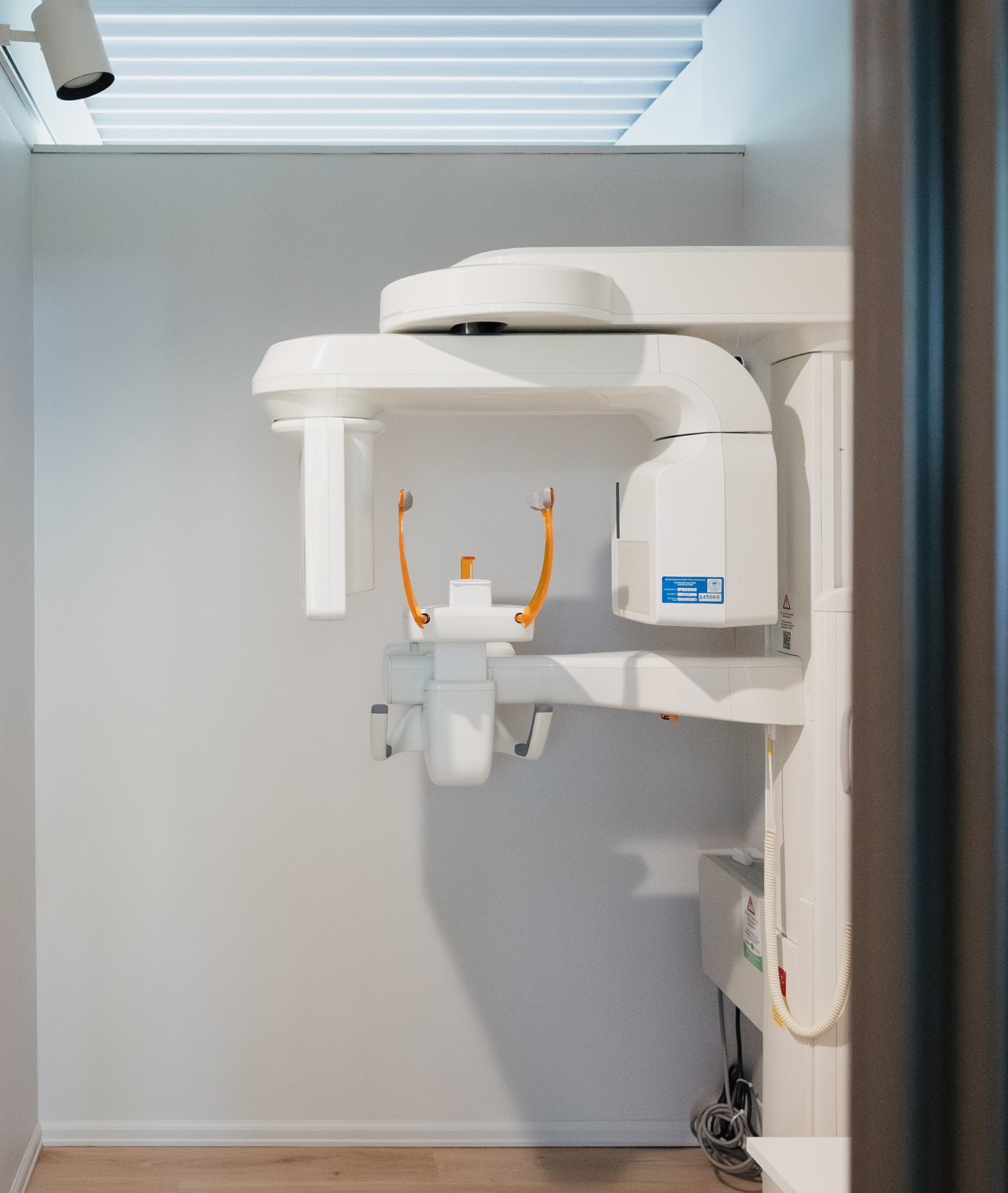 Dental X-ray machine mounted on a white wall with wooden floor below in a clean room.