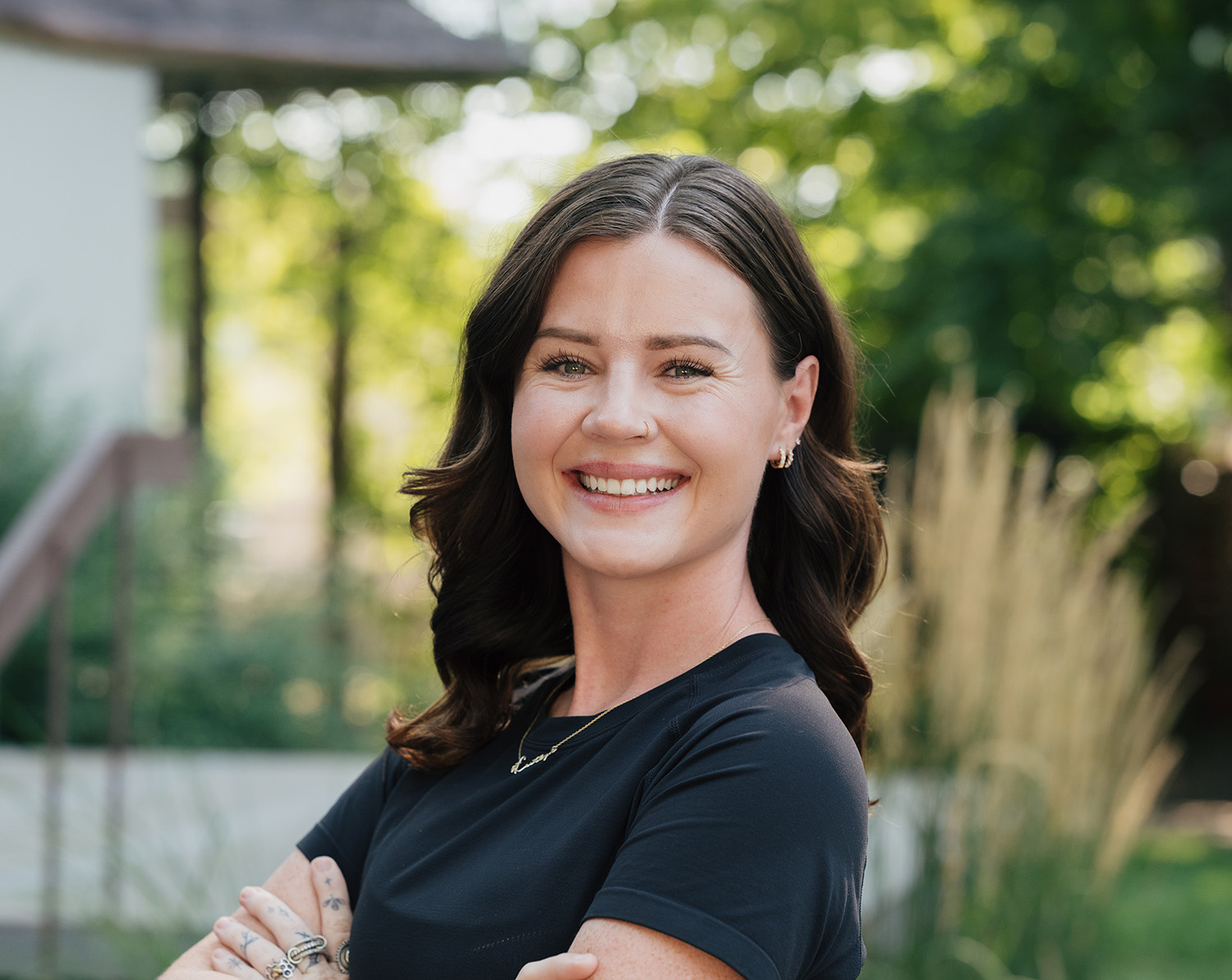 Smiling woman with dark hair, wearing a black shirt, standing outdoors with arms crossed.