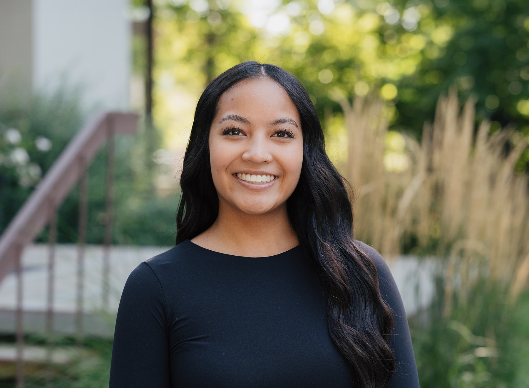 Woman with long black hair and a nose ring smiling outdoors with greenery in the background.