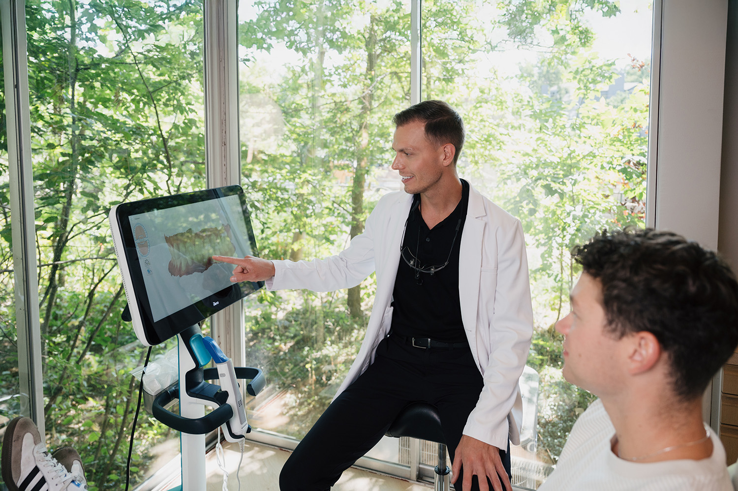 Dentist in a white coat pointing at a 3D dental scan on a screen while explaining it to a patient.