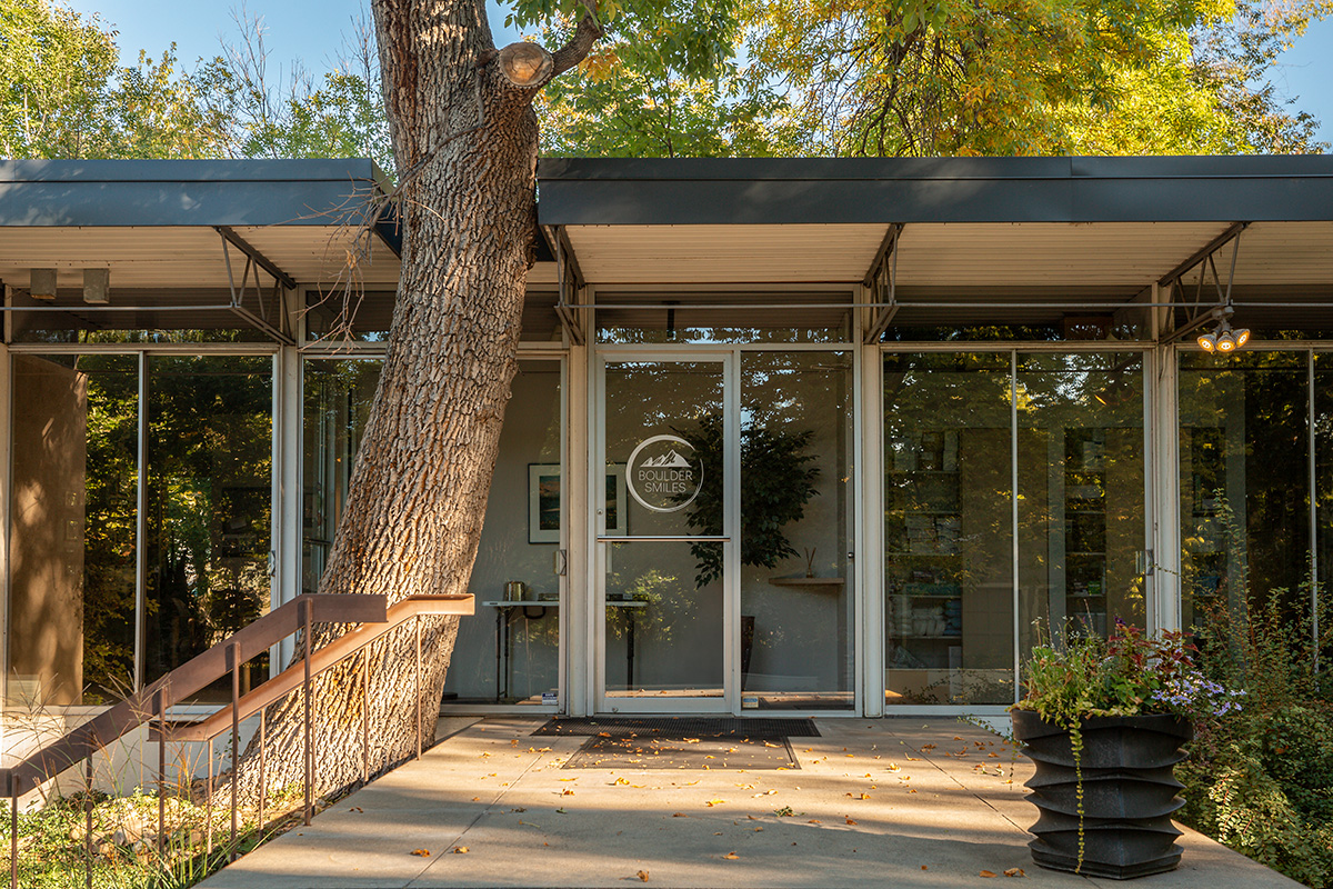 Glass entrance door of a modern building with 'Boulder Smiles' logo, surrounded by trees and plants.