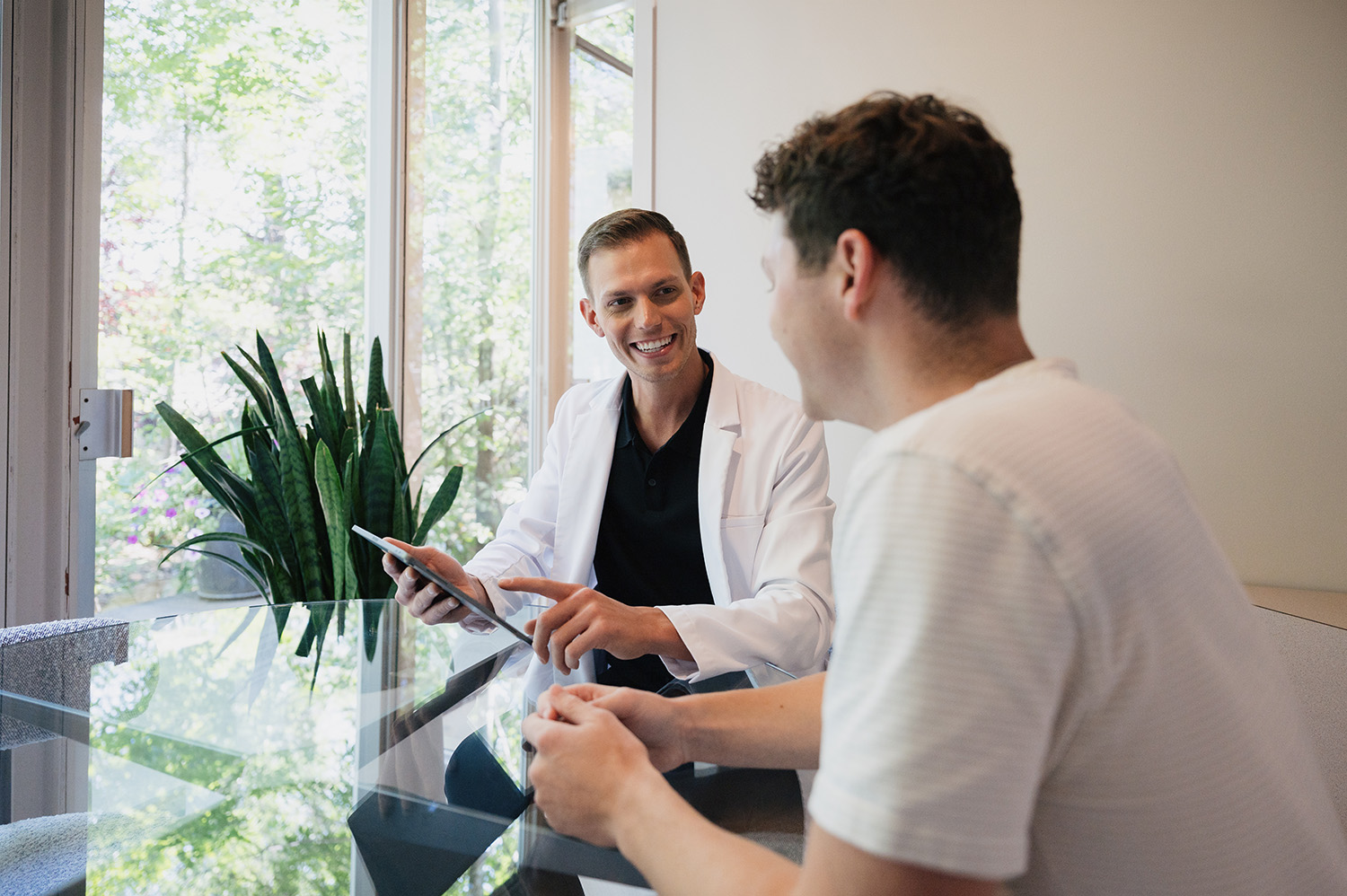 Smiling doctor in white coat shows tablet to man in white shirt at glass table by plant.