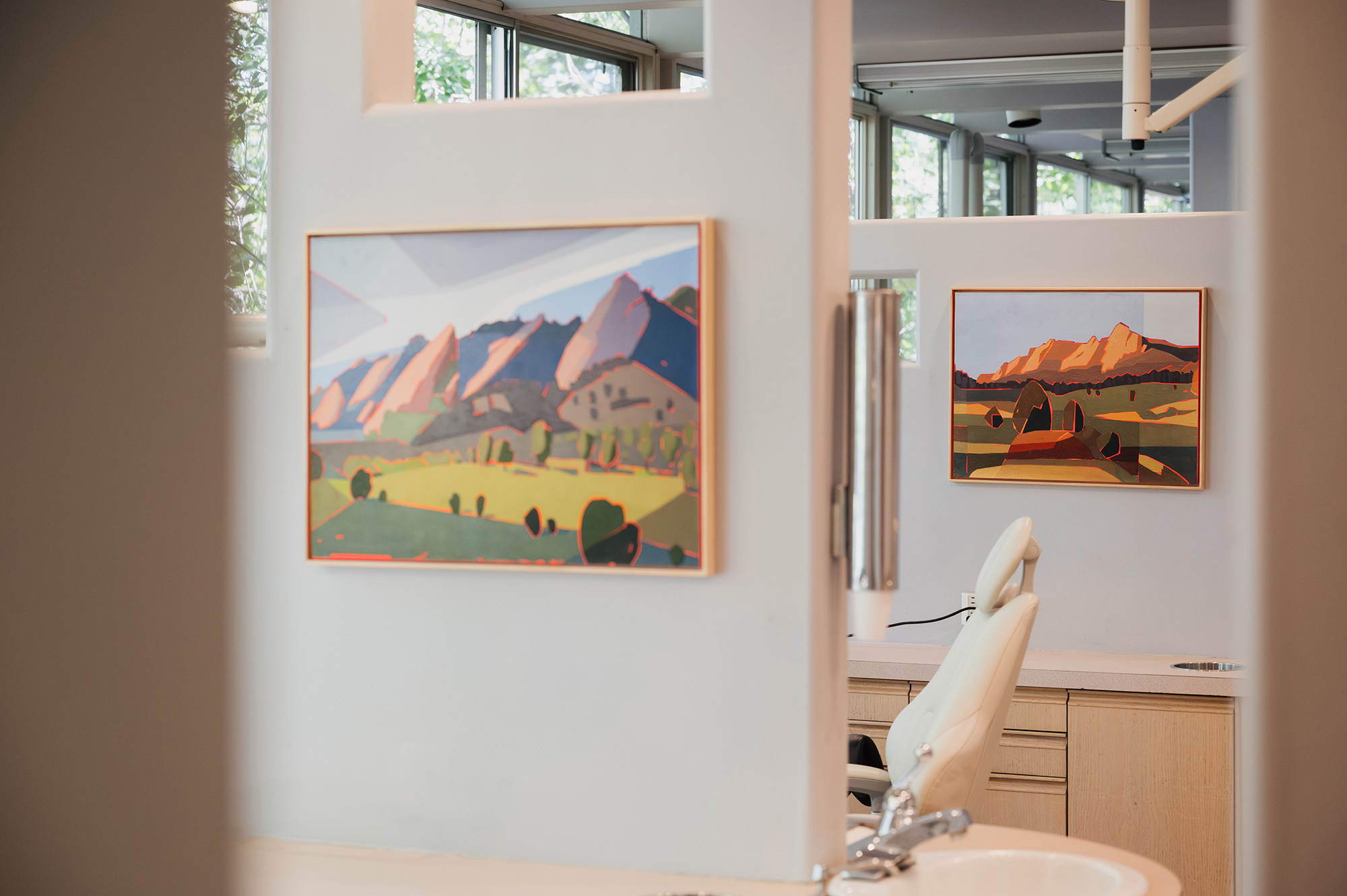 Interior of dental office with white chair, sink, and two colorful landscape paintings on walls.