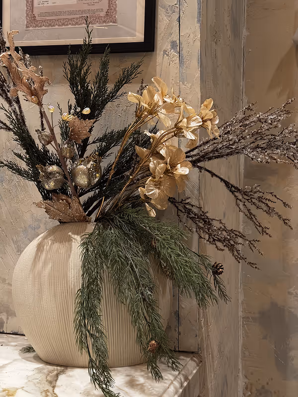 Decorative vase with a dried floral arrangement featuring golden flowers, green pine branches, and brown twigs on a marble surface against a textured beige wall.