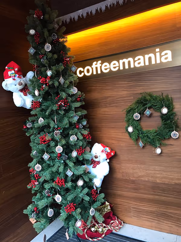 Tall Christmas tree decorated with red berries, pine cones, and hanging ornaments, two white teddy bears wearing Santa hats, and a decorated wreath hanging on a wooden wall.
