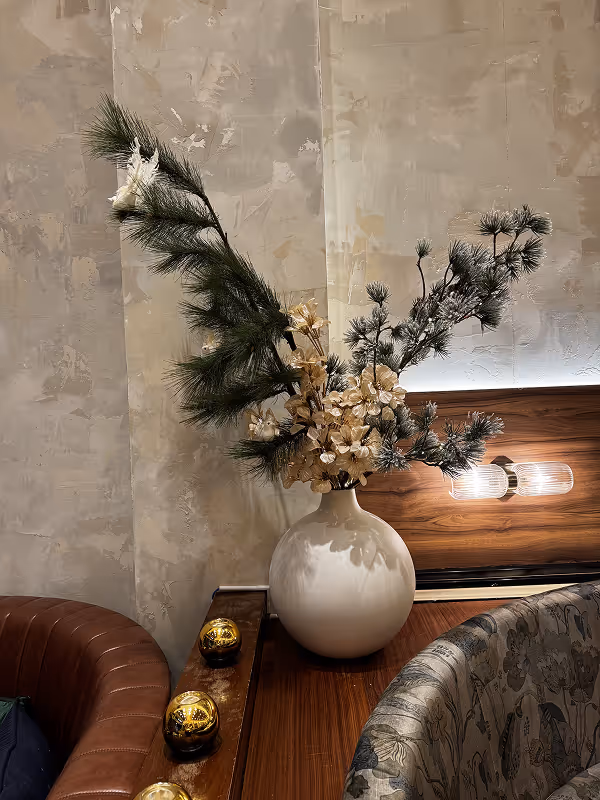 White round vase with pine branches and dried flowers on a wooden table between a brown leather chair and a floral upholstered chair.