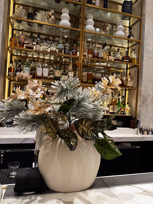 White ceramic vase with artificial snowy pine branches, beige flowers, and green leaves on a marble countertop in front of a bar shelf.
