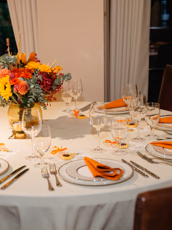 Round dining table set with white tablecloth, orange napkins, multiple wine glasses, silver cutlery, and a vibrant floral centerpiece in a glass vase.