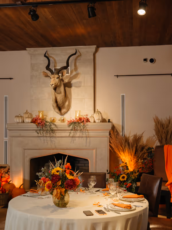 Round dining table with autumn floral centerpiece and orange napkins set for a meal in front of a decorated fireplace with a mounted kudu head.