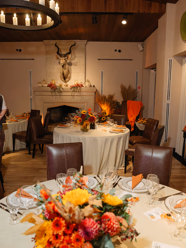 Dining room with round tables set for a meal, decorated with vibrant autumn floral centerpieces and a mounted kudu head above a fireplace.