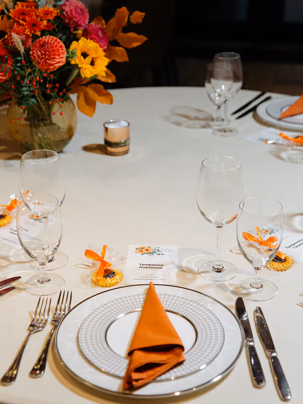 Table setting with white plates, orange napkins, clear wine glasses, silver cutlery, and a vase of vibrant orange, red, and yellow flowers.