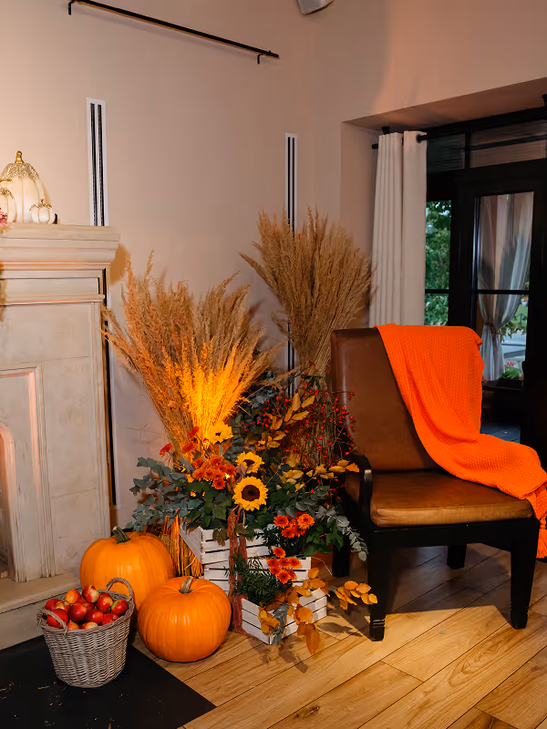 Autumn scene with two pumpkins, a basket of small red apples, an orange blanket on a brown chair, and a floral arrangement with sunflowers and dried grasses by a fireplace.