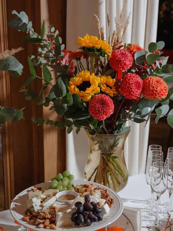 Table with a glass vase of sunflowers, pink and orange flowers, and a plate with various cheeses, grapes, nuts, and honey.