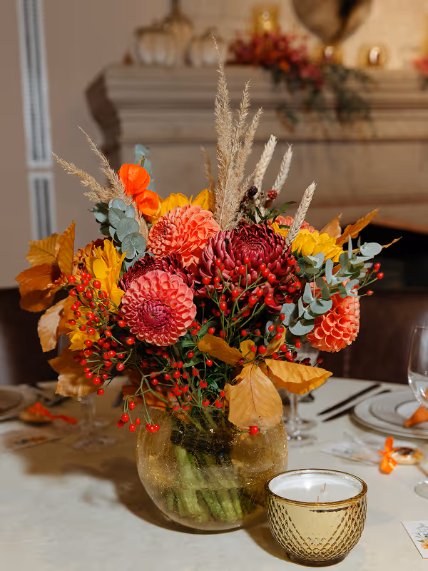 Vase with vibrant autumn floral arrangement of red, orange, and yellow flowers and berries on a table beside a candle in a gold holder.