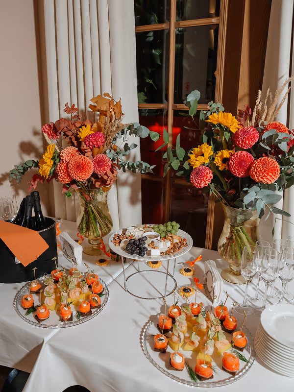 Table set with two glass vases of colorful autumn flowers, cheese and grape platter on a stand, trays of smoked salmon and shrimp appetizers, stacked white plates, and empty wine glasses.