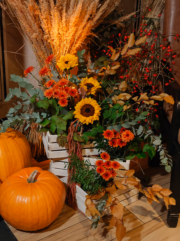 Autumn arrangement with sunflowers, orange flowers, dried grasses, red berries, and two pumpkins on a wooden floor.