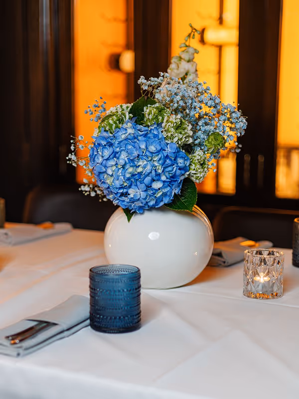 White vase with blue and white flowers on a white tablecloth, surrounded by a blue glass and a lit candle in a glass holder.
