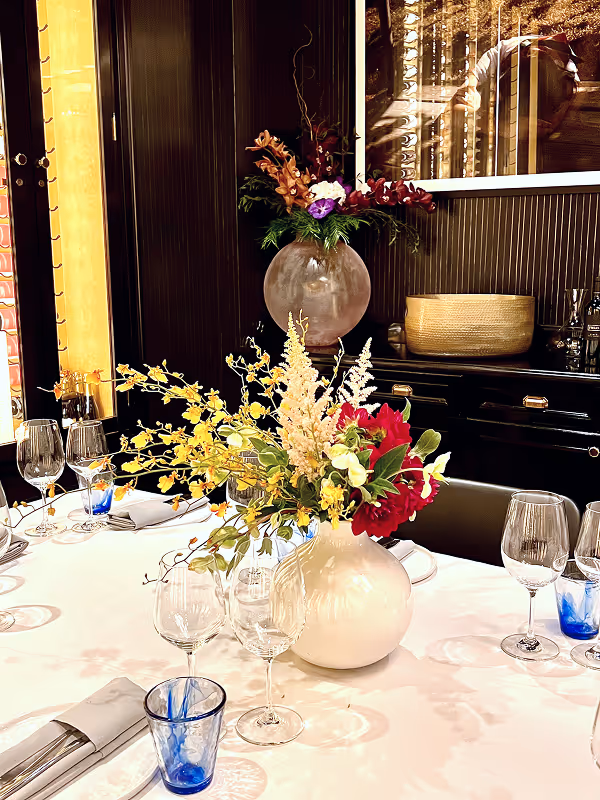 Dining table set with white tablecloth, clear and blue glassware, and a floral centerpiece featuring red, yellow, and white flowers in a white vase.
