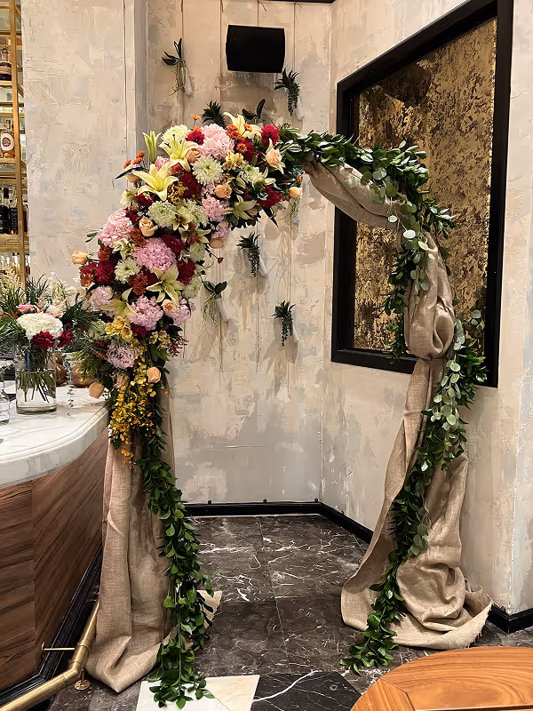 Decorative arch adorned with a variety of colorful flowers and green foliage, draped with beige fabric, standing indoors near a marble floor and a wooden bar counter.