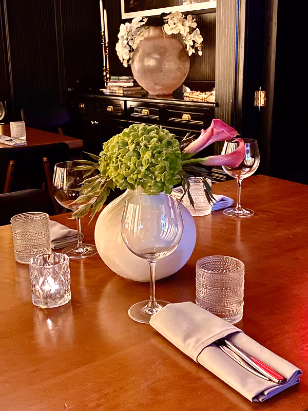 Wooden table set with a white rounded vase holding green hydrangea and pink calla lilies, surrounded by empty wine glasses, candles, and neatly folded napkins with silverware.