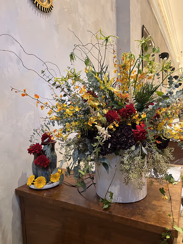 Large floral arrangement with red, yellow, and green flowers and foliage in a white pot on a wooden table next to a smaller vase with red and yellow flowers.