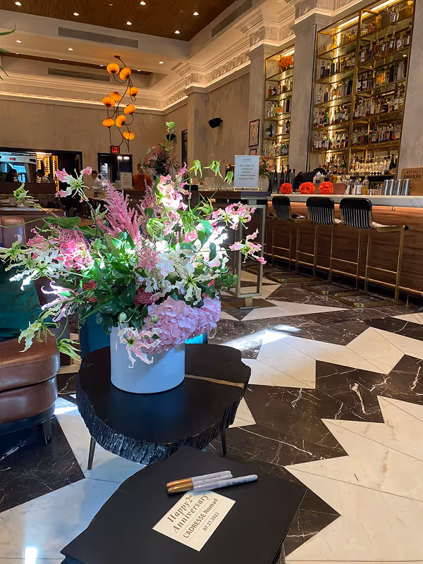 Large bouquet of pink and white flowers in a white vase on a black table in a restaurant with marble floors and wooden furniture.