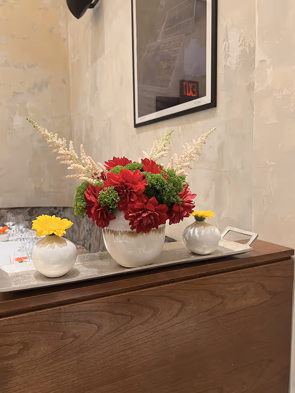 Table centerpiece with a white vase of red flowers and green foliage flanked by two smaller white vases each holding a single yellow flower.