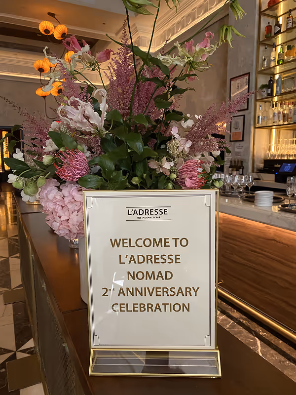Floral arrangement with pink and purple flowers behind a sign welcoming guests to the restaurant bar.