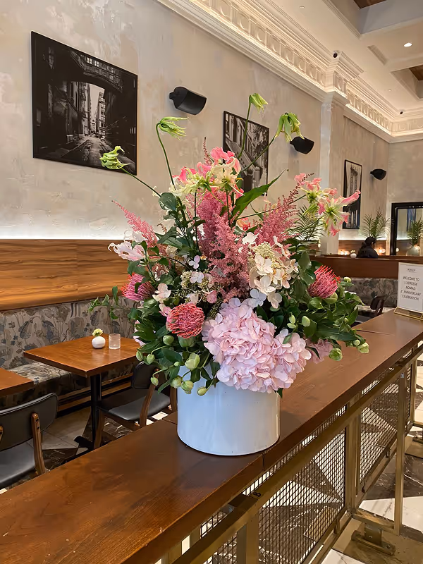 Large floral arrangement with pink and white flowers in a white vase on a wooden counter inside a restaurant.
