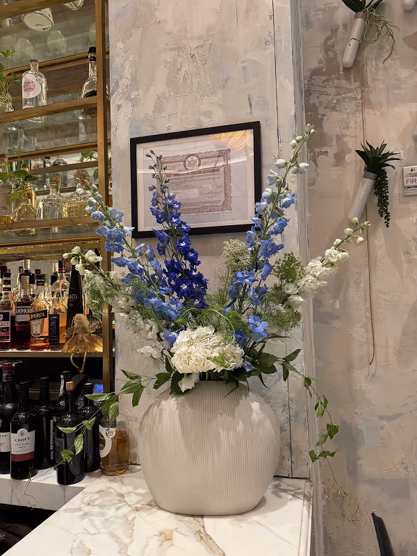 Large ceramic vase with white and blue flowers arranged on a marble countertop beside a bar and a framed certificate on a textured wall.
