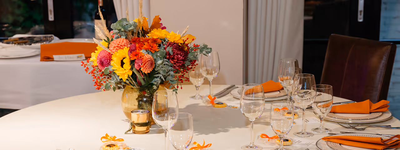 Round dining table set with white tablecloth, orange napkins, glassware, silverware, and a colorful floral centerpiece.