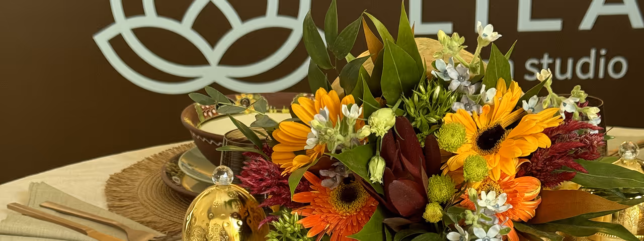 Table set with fall-themed centerpiece of orange, yellow flowers and green leaves, brown glass tumbler, beige candle with black band, and autumn decorations on woven placemats.