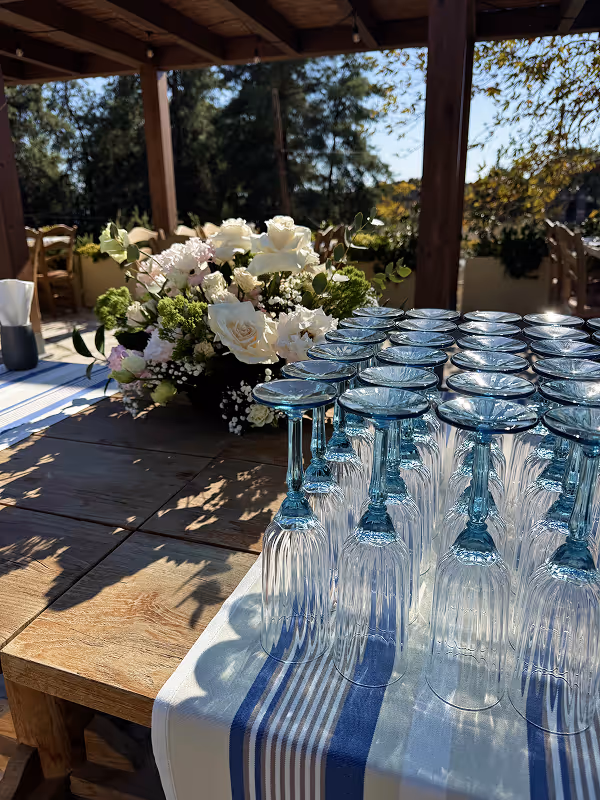 Rows of upside-down blue-tinted wine glasses on a striped cloth-covered wooden table with a white and green floral arrangement in an outdoor shaded area.