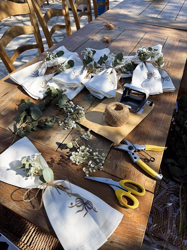 Wooden table with white napkins tied with twine, decorated with small white flowers and eucalyptus leaves, scissors, pruning shears, twine spool, and a hole punch.