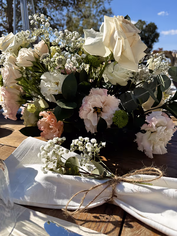 Bouquet of white, pink, and green flowers on a wooden table, next to a white napkin tied with twine and decorated with small white flowers.