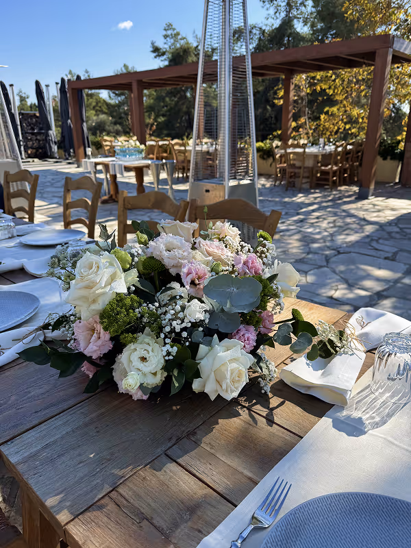 Outdoor wooden table set for dining with a centerpiece of white and pink flowers and greenery under clear blue sky.