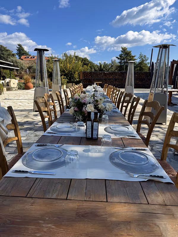 Long outdoor wooden dining table set with glass plates, cutlery, empty glasses, flower centerpiece, and bottles under a blue sky with scattered clouds.